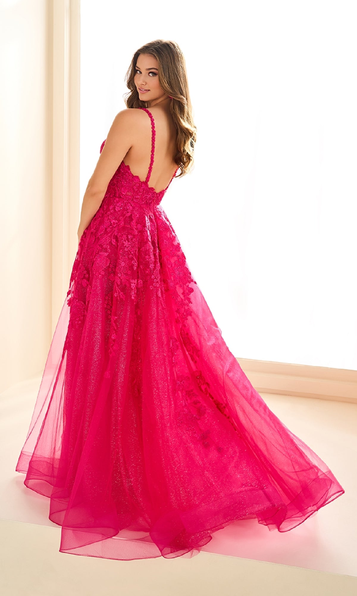 A woman stands in a brightly lit room wearing the Ellie Wilde EW36015 long prom dress, featuring bright pink glitter tulle, a floor-length flowing skirt, floral appliqués, and a corset bodice as she looks over her shoulder at the camera.